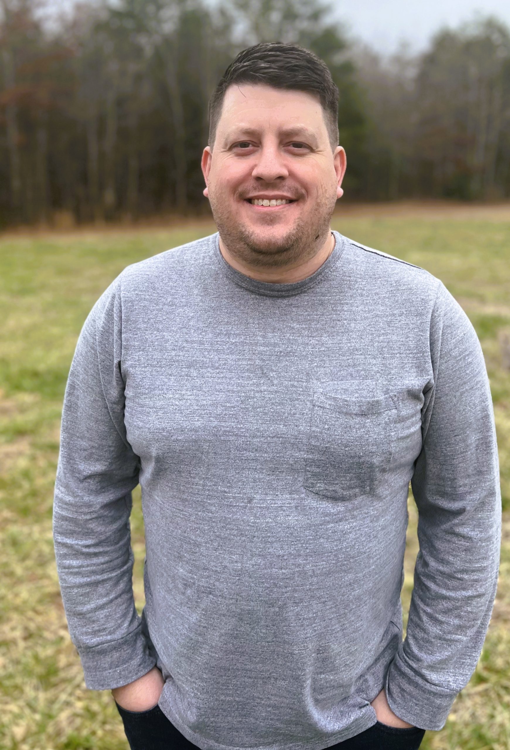 Kyle in a gray shirt standing in with grass and trees behind him.
